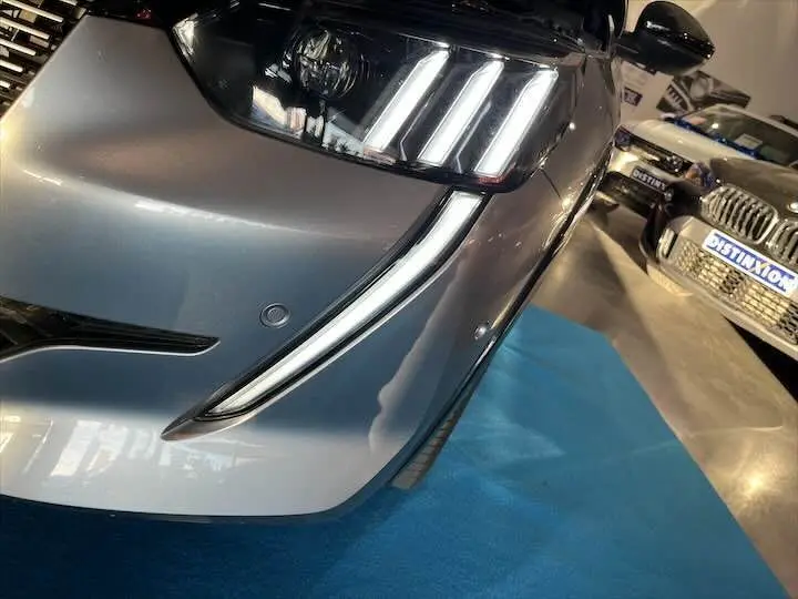 Gros plan sur le phare avant droit et la calandre d'une Peugeot 208 E-208 GT gris Artense, éclairage LED distinctif visible.