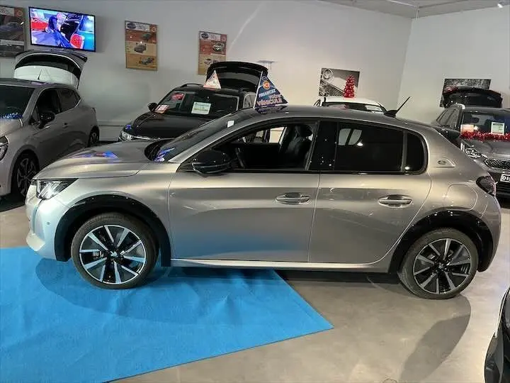 Peugeot e-208 gris Artense vue de profil côté gauche, exposée en intérieur sur tapis bleu.