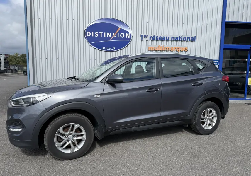 Vue de profil côté gauche d'un Hyundai Tucson gris 2015 garé devant un bâtiment avec logo Distinxion.