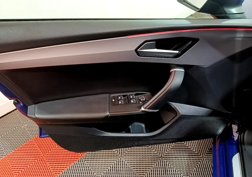 Vue intérieure côté gauche de la porte avant d'une SEAT Leon bleu, avec poignée cuir et surpiqûres rouges.