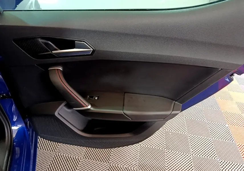 Vue rapprochée de la porte avant gauche bleu de la SEAT LEON 1.5 eTSI avec poignée noire et surpiqûres rouges.
