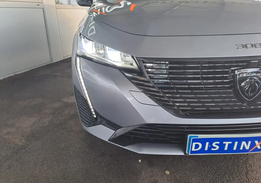 Vue de face côté gauche d'une Peugeot 308 gris Artense 2023 avec phares LED allumés et calandre noire distinctive.