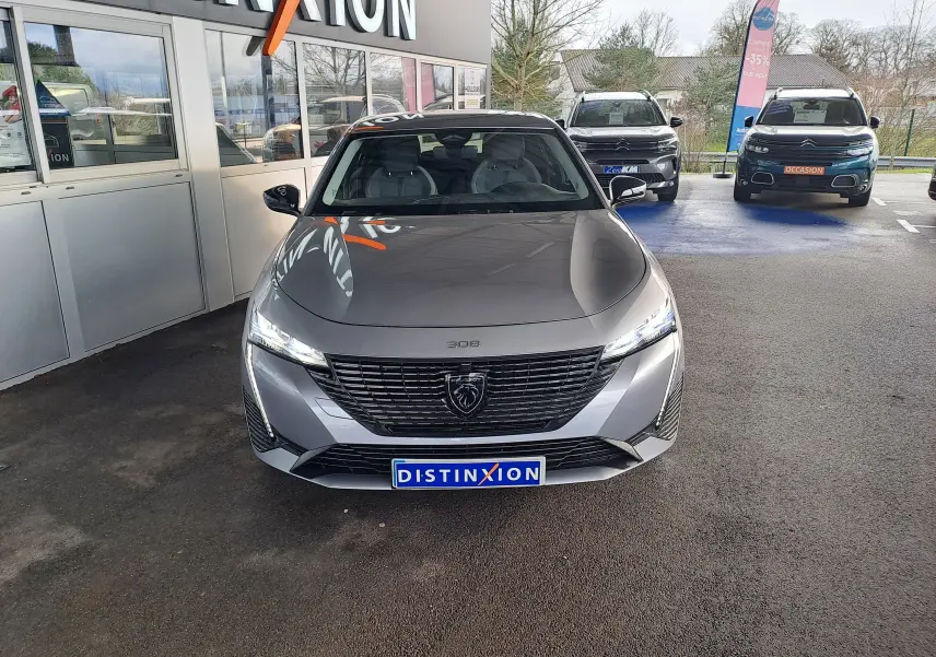 Peugeot 308 gris Artense vue de face, avec calandre noire et phares LED allumés, stationnée devant un showroom.