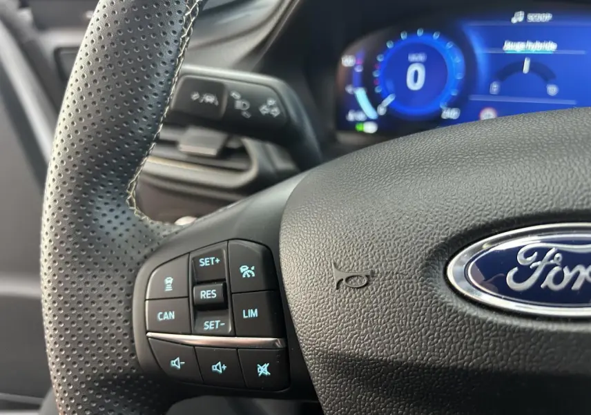 Gros plan sur le volant perforé noir du Ford Puma avec commandes intégrées et tableau de bord numérique bleu en arrière-plan.