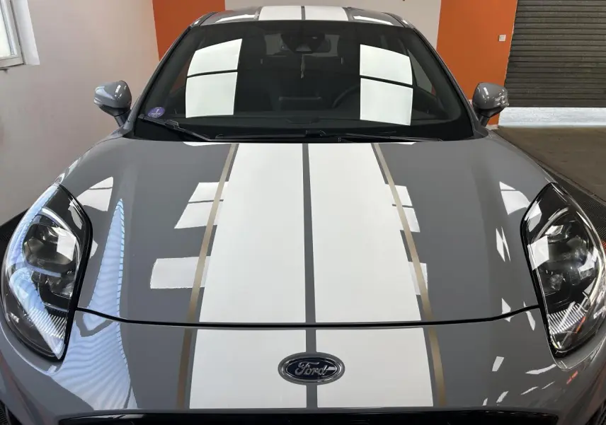 Vue avant rapprochée du Ford Puma gris avec bandes blanches et jantes ST Line Gold visibles en reflet.