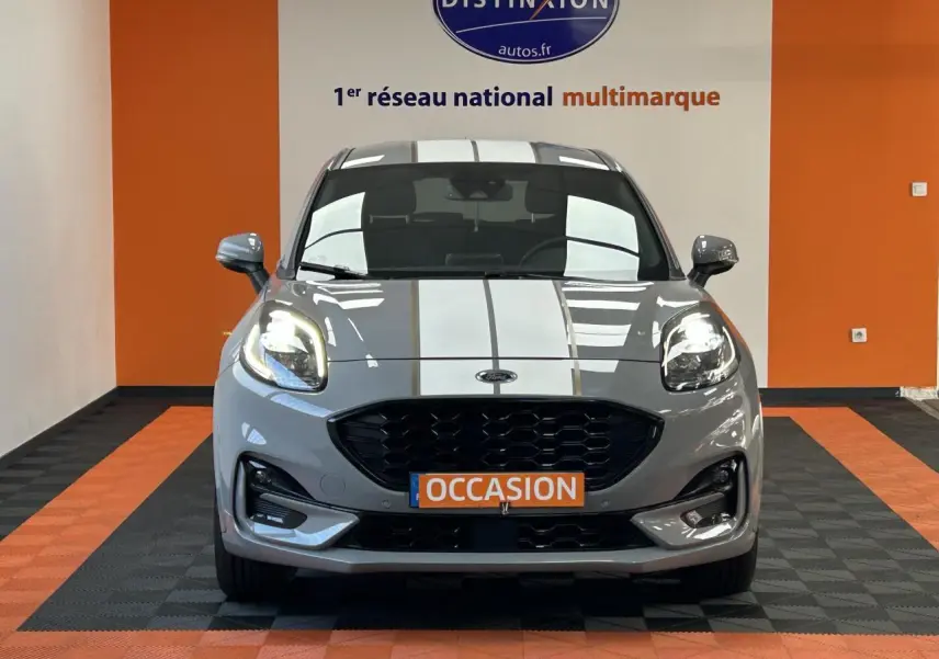 Vue frontale d'un Ford Puma gris avec bandes blanches, phares allumés et plaque "OCCASION" orange.