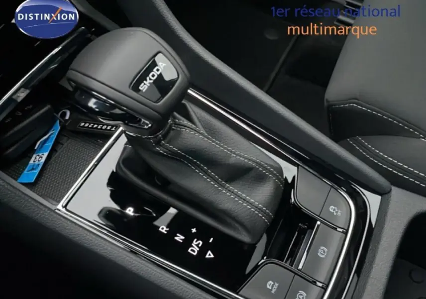 Gros plan sur le levier de vitesses automatique noir avec logo Skoda, entouré de commandes et coutures blanches.