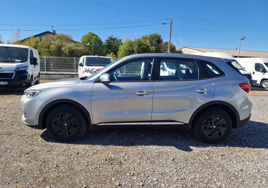 Vue latérale droite d'un MG Motor 1.5 L Hybrid+ 2026 en Cosmic Silver avec jantes noires sur un sol gravillonné.