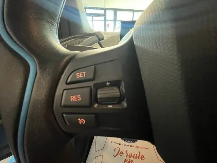 Gros plan sur les commandes de régulateur de vitesse au volant d'une BMW i3 noire, intérieur gris-bleu visible en arrière-plan.