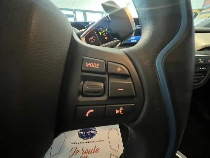 Gros plan sur les commandes audio et vocales au volant d’une BMW i3 noire, avec affichage digital en arrière-plan.