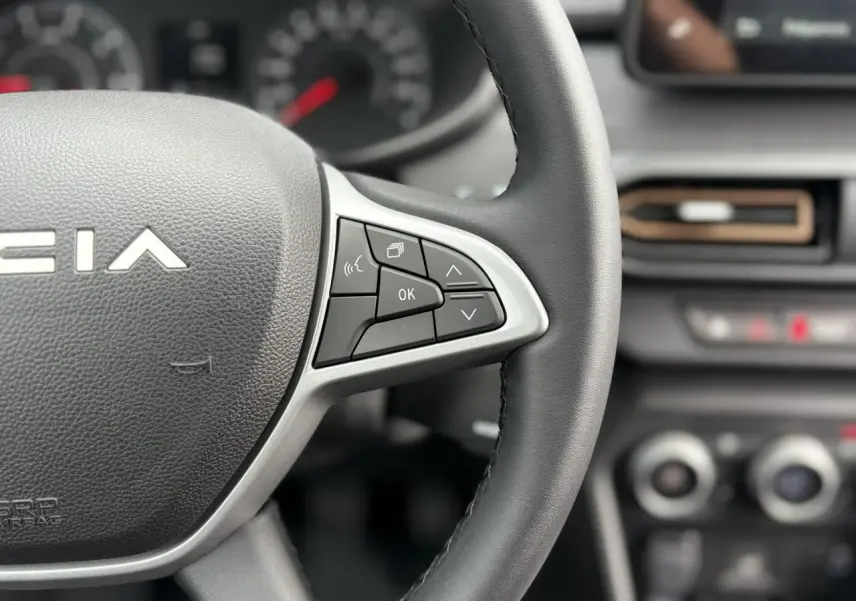Gros plan sur le volant noir de la Dacia Jogger 2025 avec commandes multifonctions et tableau de bord flou en arrière-plan.