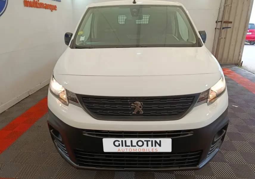 Vue avant d'un Peugeot Partner blanc avec calandre noire et phares allumés dans un garage.