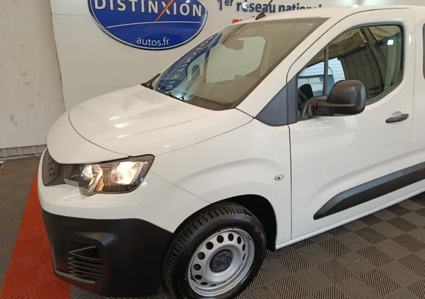 Vue 3/4 avant gauche d’un Peugeot Partner blanc avec pare-chocs noir et jante acier simple en intérieur showroom.