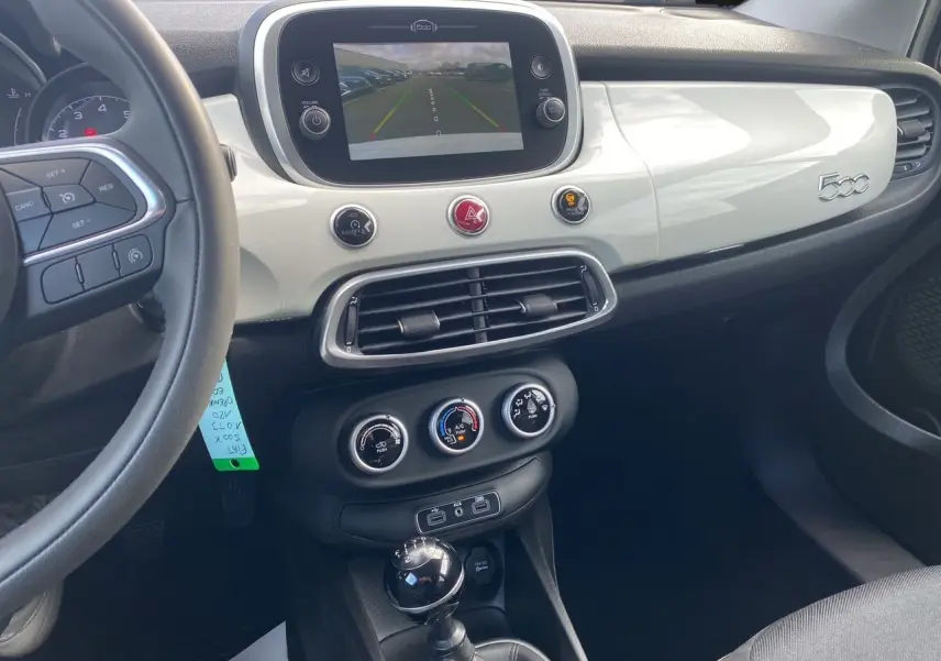 Vue intérieure du tableau de bord blanc brillant et console centrale noire d'une Fiat 500X avec écran tactile et levier de vitesse manuel.