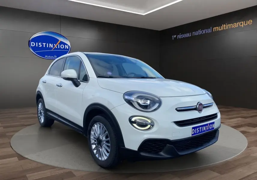 Fiat 500X blanc en 3/4 avant droit, avec phares LED allumés et jantes alliage 17 pouces dans un showroom.