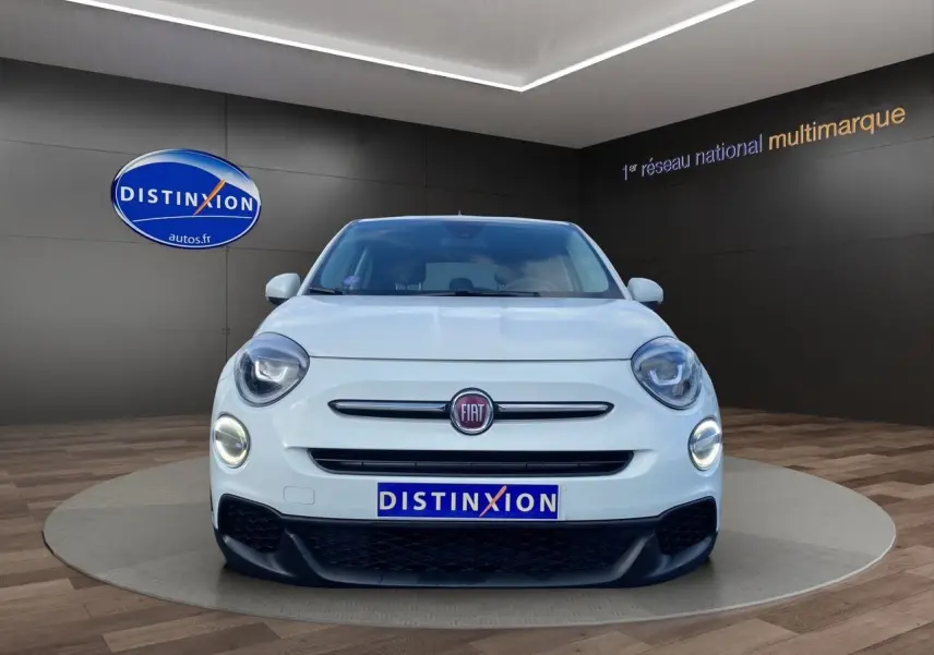 Vue frontale d'une Fiat 500X blanche avec phares allumés et logo FIAT bien visible sur un fond intérieur sombre.