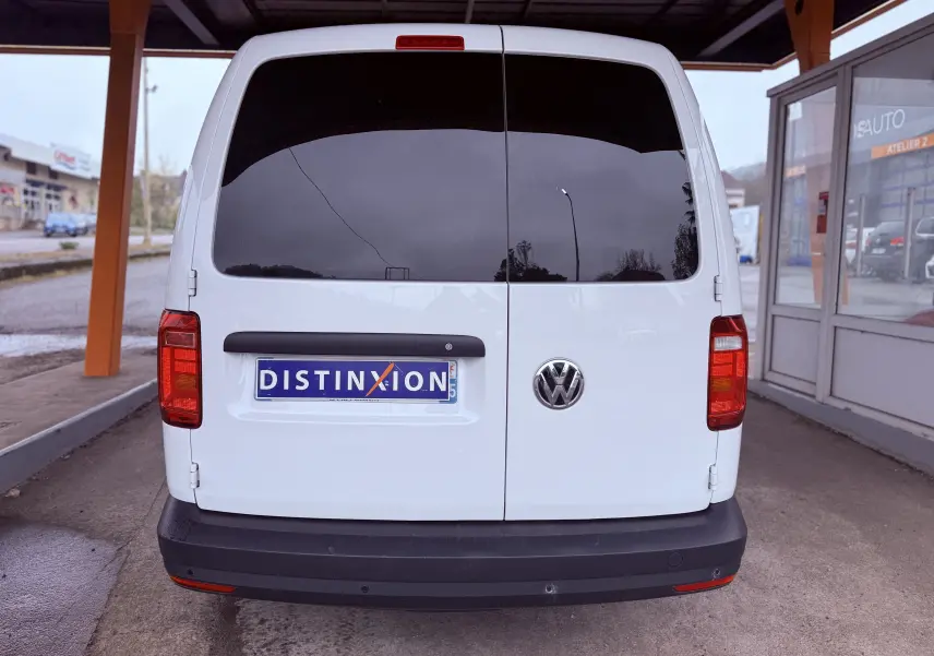 Vue arrière d'un Volkswagen Caddy Maxi Long blanc garé sous abri, avec vitres arrière teintées et plaque Distinxion.