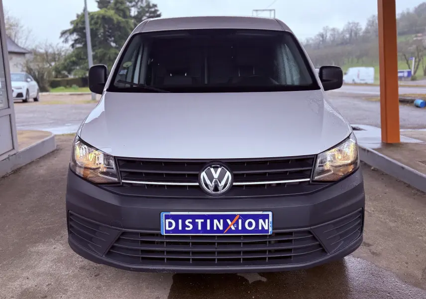 Vue de face d'un Volkswagen Caddy blanc avec pare-chocs noir et phares allumés sous abri.