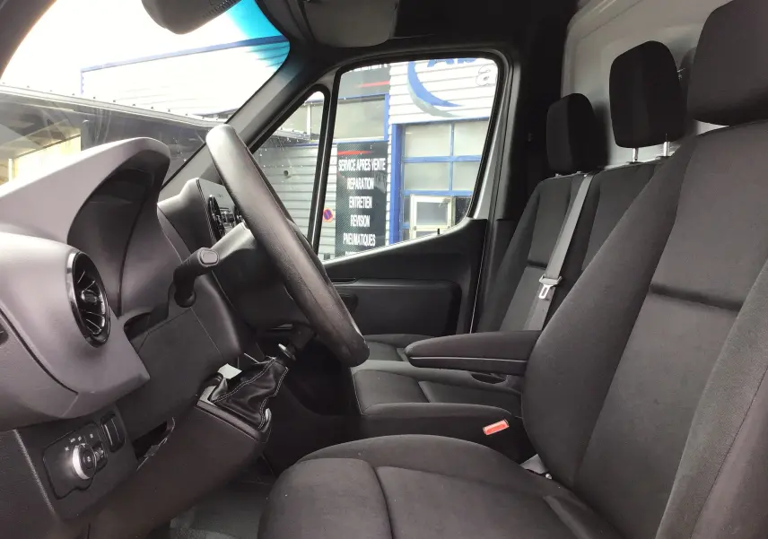 Vue intérieure côté conducteur du Mercedes Sprinter blanc 2018, montrant le volant, levier de vitesses manuel et sièges en tissu gris.