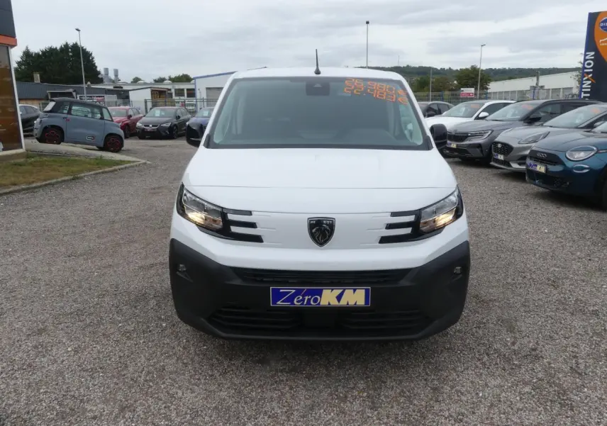 Vue de face d'un Peugeot Partner blanc avec calandre noire et logo lion, stationné sur un parking extérieur.