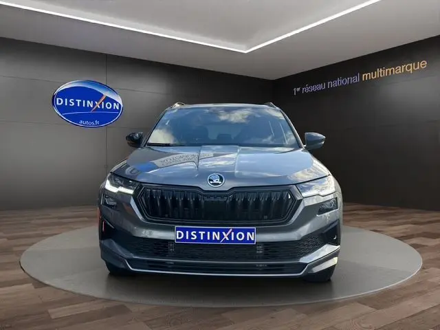 Vue de face d'un Skoda Karoq gris graphite 2025 avec calandre noire et feux LED allumés sur fond intérieur showroom.