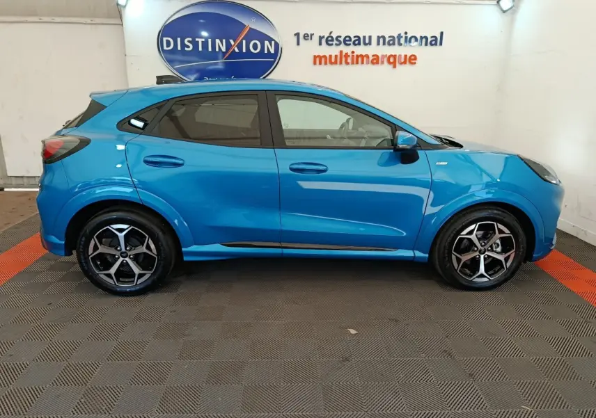 Vue latérale gauche d'un Ford Puma bleu 2025 ST-LINE avec jantes alliage et vitres teintées en intérieur showroom.