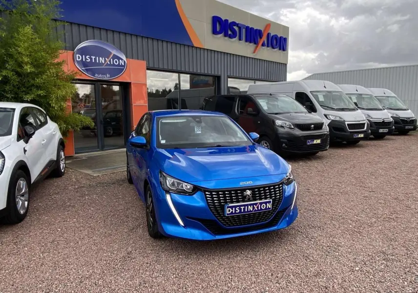 Peugeot 208 bleu en 3/4 avant stationnée devant un concessionnaire avec plusieurs véhicules utilitaires en arrière-plan.
