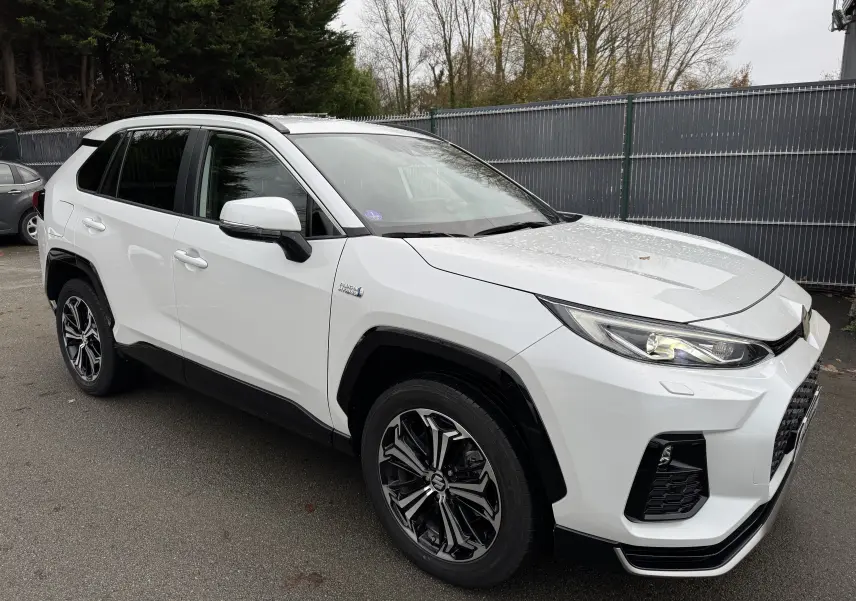 Suzuki Across blanc en vue 3/4 avant droit, avec phares allumés et jantes alliage noires distinctives.