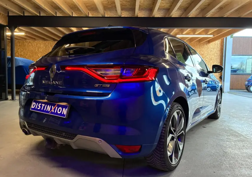 Vue 3/4 arrière droite d'une Renault Mégane GT Line bleu métallisé, avec feux arrière allumés, stationnée en garage.