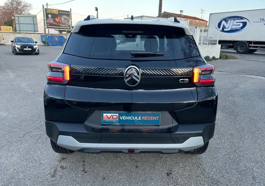 Vue arrière d'une Citroën C3 noire 2025 avec barres de toit et feux arrière allumés sur parking.