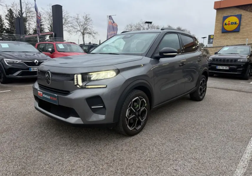 Citroën C3 gris vue 3/4 avant droit, phares allumés, jantes alliage noires et barres de toit visibles.