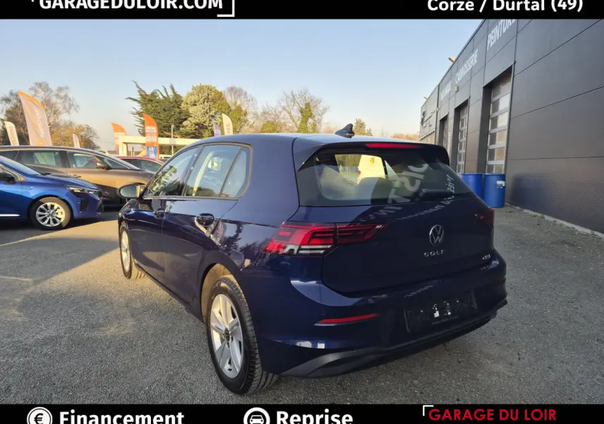 Vue 3/4 arrière droite d'une Volkswagen Golf bleue 2023 stationnée devant un garage, avec feux arrière LED visibles.
