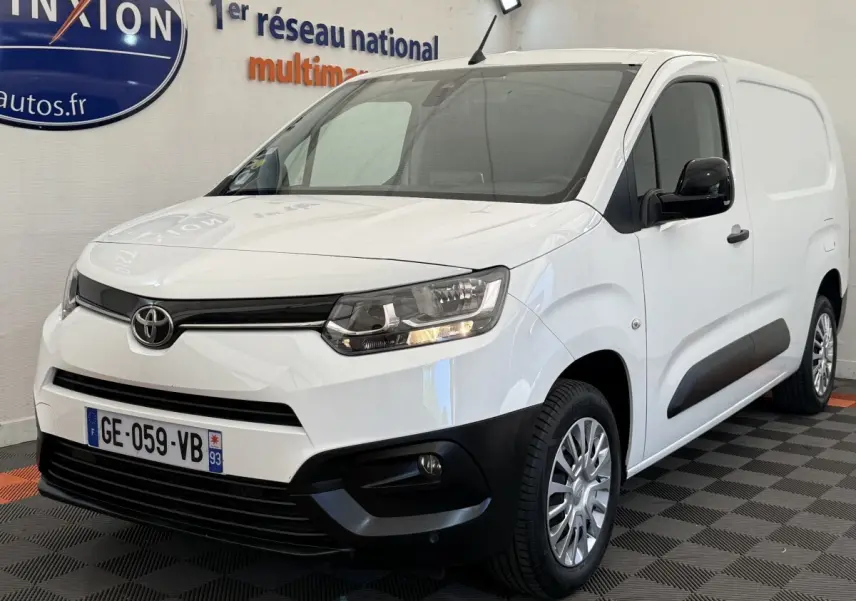 Toyota Proace City blanc en vue 3/4 avant droit, avec calandre noire et phares allumés dans un showroom.