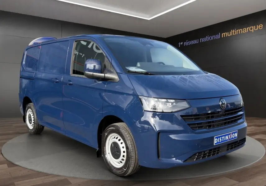 Vue 3/4 avant d’un Volkswagen Transporter bleu avec jantes acier et calandre noire, en intérieur showroom.