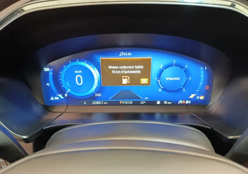 Tableau de bord numérique bleu du Ford Kuga 2024 affichant une alerte de faible carburant, vu de face depuis le volant.