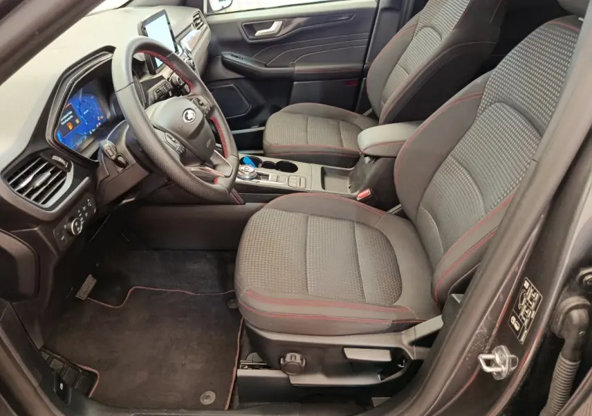 Vue intérieure côté conducteur du Ford Kuga 2024 St Line, sièges tissu gris avec surpiqûres rouges et tableau de bord digital.