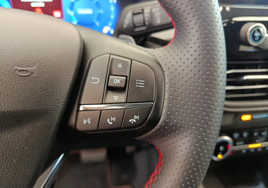 Gros plan sur le volant en cuir perforé avec surpiqûres rouges du Ford Kuga 2024, commandes multifonctions visibles.