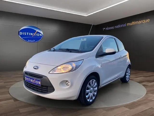 Vue 3/4 avant droite d'une Ford KA blanche de 2015 avec phares allumés, sur fond intérieur showroom.