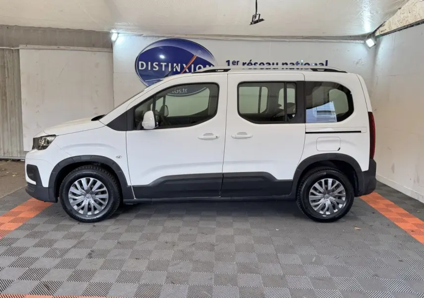 Vue de profil côté gauche d'un Peugeot Rifter blanc 2019 avec protections noires et jantes argentées en intérieur showroom.