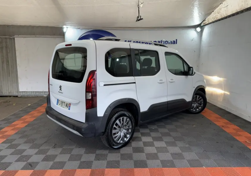 Vue 3/4 arrière droite du Peugeot Rifter blanc avec barres de toit et protections latérales noires dans un garage.