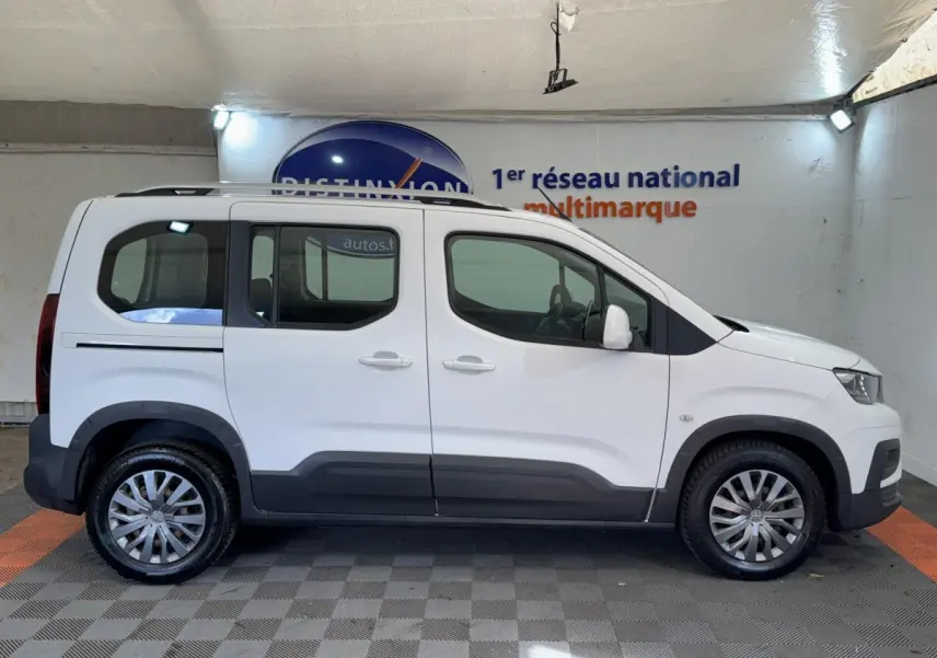 Profil droit d'un Peugeot Rifter blanc 2019 avec jantes alliage et barres de toit chromées en intérieur showroom.