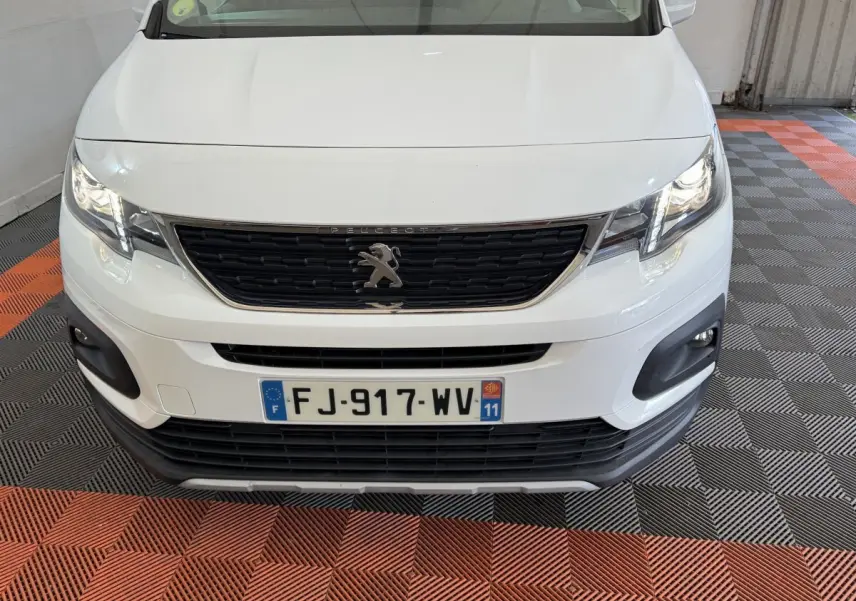 Vue avant rapprochée d’un Peugeot Rifter blanc avec calandre noire et phares allumés dans un garage.