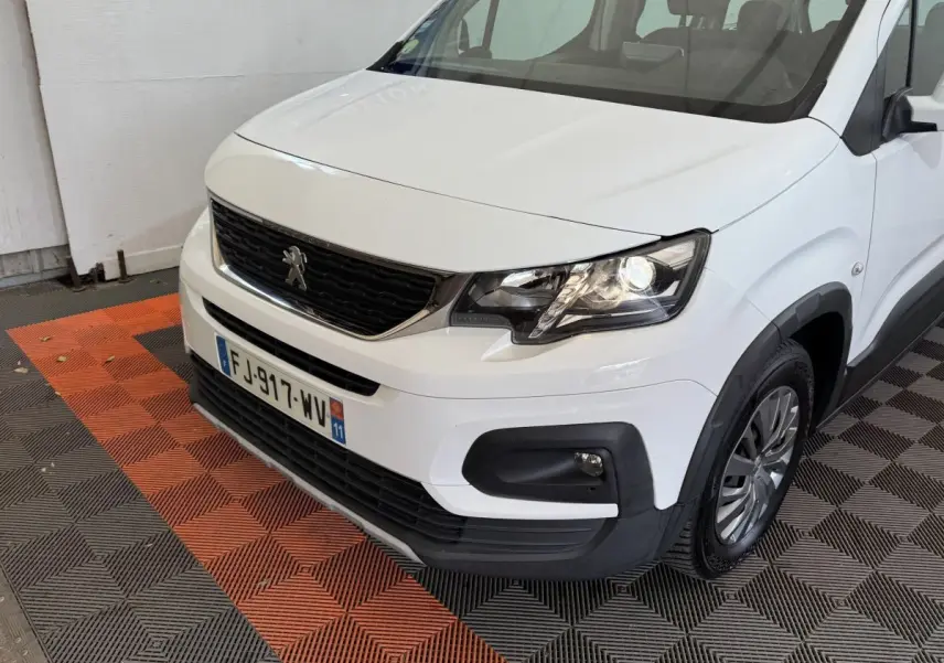 Vue 3/4 avant droit d'un Peugeot Rifter BlueHDi 100 Allure blanc avec calandre noire et feux allumés.