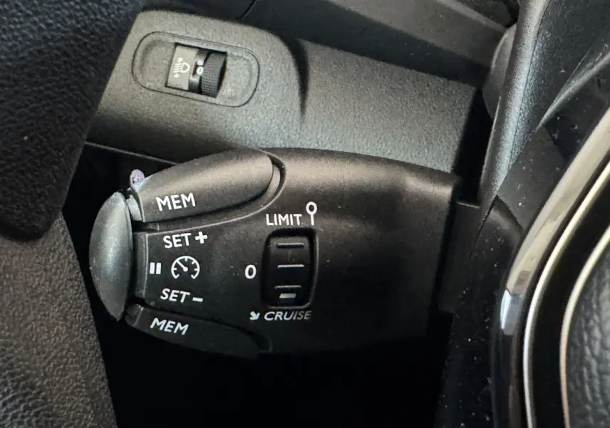 Gros plan sur la commande de régulateur et limiteur de vitesse noire dans l'habitacle du Peugeot Rifter blanc.