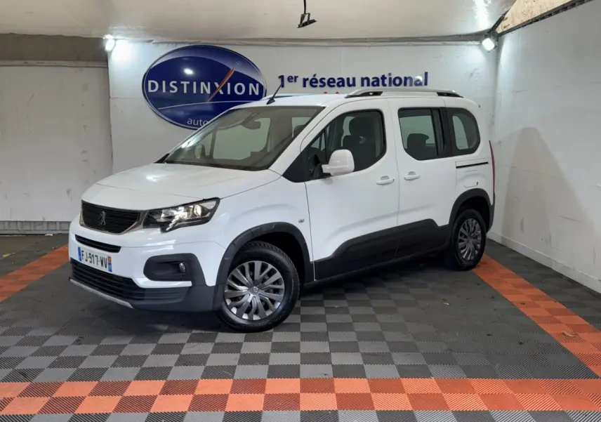 Peugeot Rifter blanc en 3/4 avant droit, avec barres de toit noires et protections latérales noires visibles.