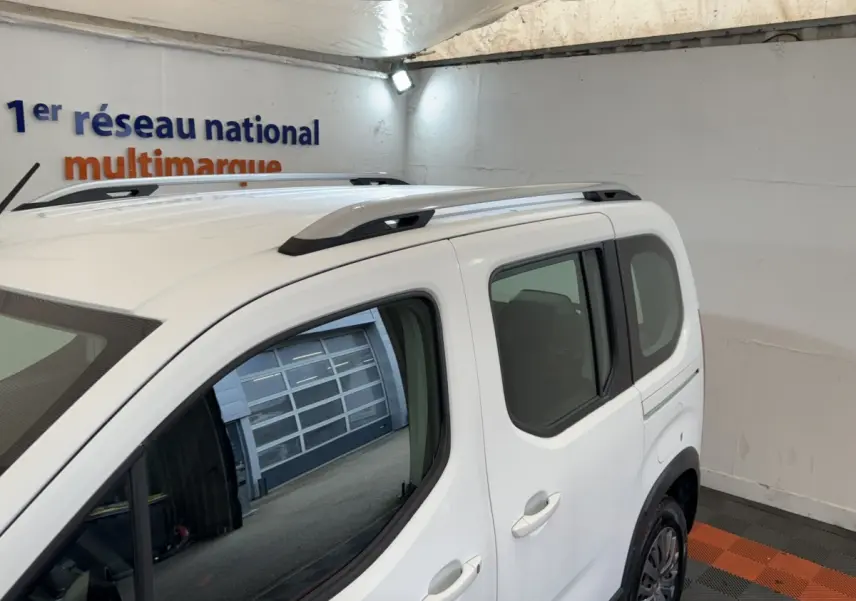 Vue 3/4 arrière droite du Peugeot Rifter blanc avec barres de toit noires et vitres teintées dans un garage.