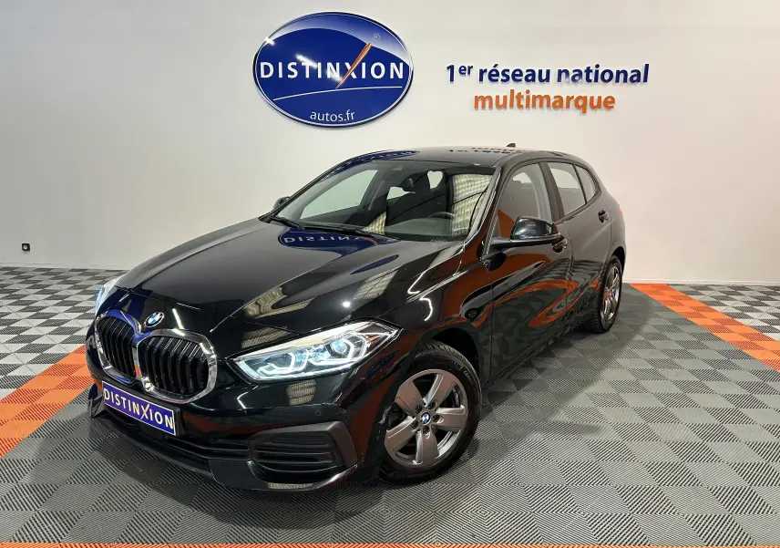BMW Série 1 16d noire vue en 3/4 avant droit, phares LED allumés, sur sol damier gris et orange en showroom