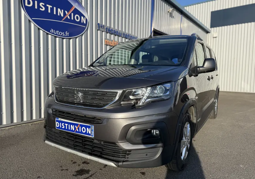 Peugeot Rifter gris platinium en 3/4 avant droit, avec calandre noire et jantes alliage visibles, stationné devant un bâtiment.