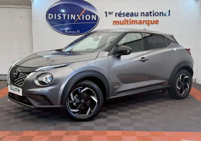 Nissan Juke gris foncé en 3/4 avant droit, avec jantes noires et logo Hybrid visible sur l'aile avant.