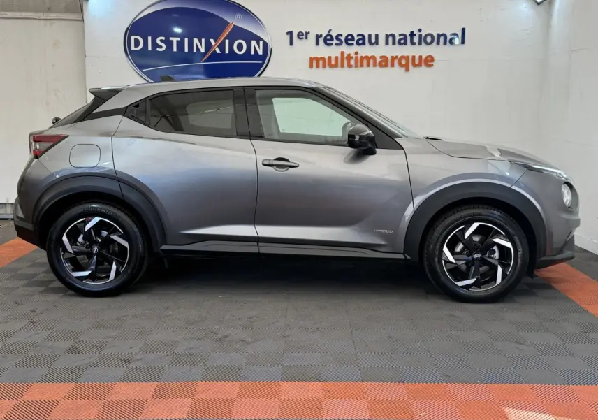 Vue de profil côté gauche d'un Nissan Juke gris foncé 2023 hybride avec jantes noires et logo Distinxion en arrière-plan.